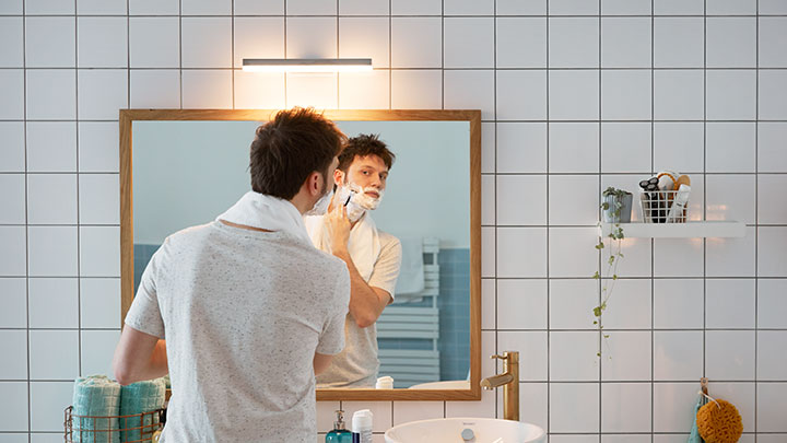 Mand barberer sig foran et badeværelsesspejl oplyst af en vægmonteret Philips LED pære